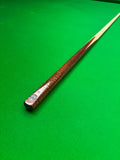 A close-up of a wooden snooker cue shaft with a polished brown and amber patterned handle, resting diagonally on a green felt pool table.