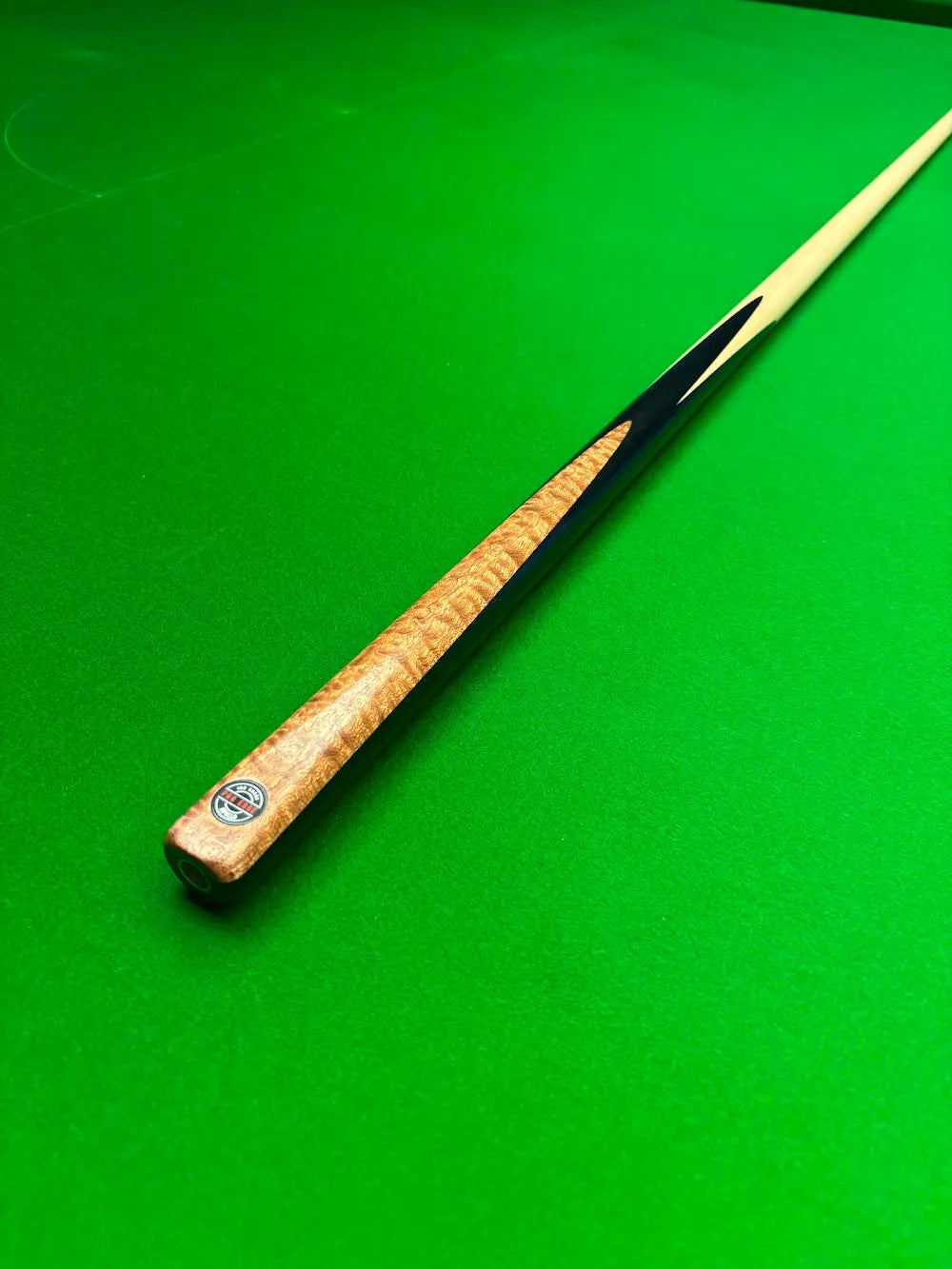 A close-up of a snooker cue shaft resting diagonally on a vibrant green felt table surface, showing the natural wood grain handle and a black ferrule.