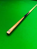 A close-up of a snooker cue shaft resting diagonally on a vibrant green felt table surface, showing the natural wood grain handle and a black ferrule.