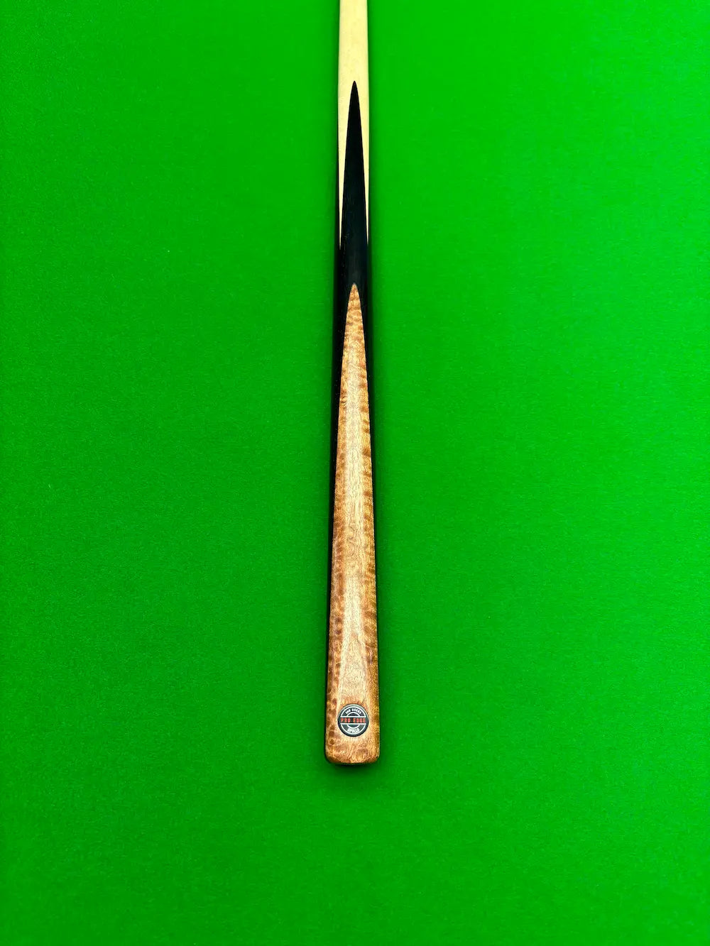 A close-up of a snooker cue shaft and butt end against a bright green felt background. The shaft is light wood with a black ferrule, and the butt cap features a circular black and red Pro Edge logo.