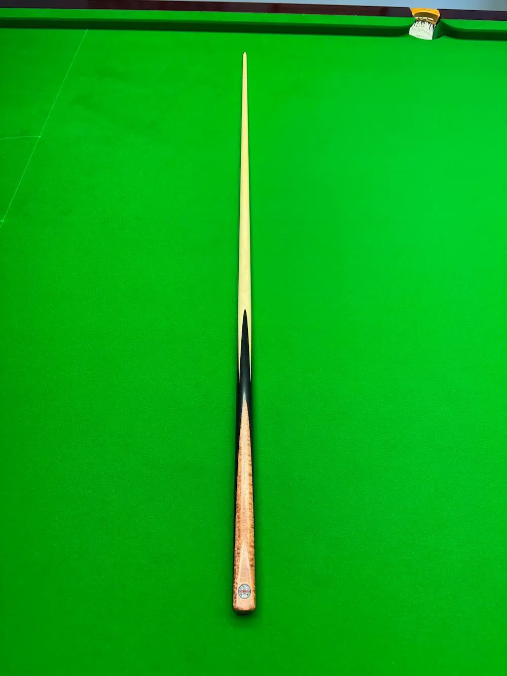 A single snooker cue laid vertically on a bright green snooker table, with a wooden handguard and a light-colored shaft.