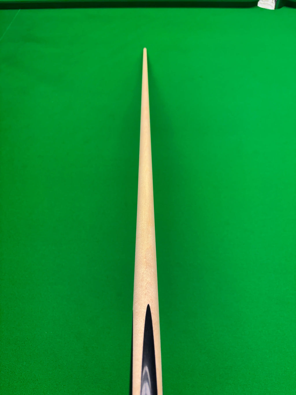 A close-up, top-down view of a wooden cue shaft pointing vertically down the center of a green snooker table.