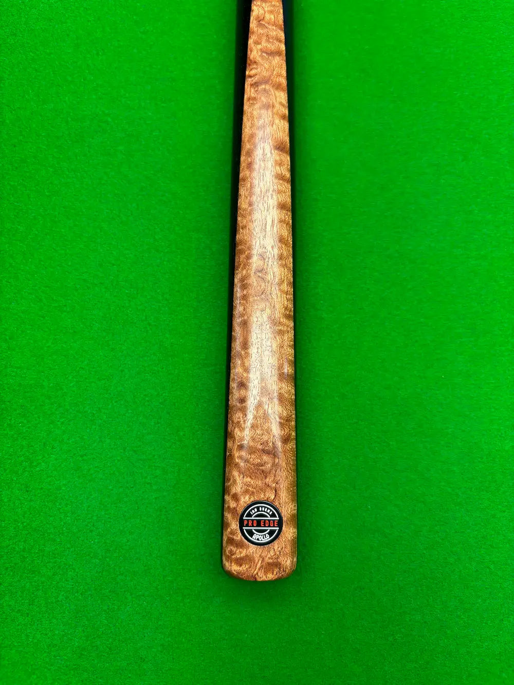 A close-up of a wooden snooker cue shaft with a prominent figured wood grain pattern, featuring a circular black and white 'Pro Edge' brand logo near the butt, set against a bright green felt background.
