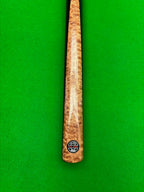 A close-up of a wooden snooker cue shaft with a prominent figured wood grain pattern, featuring a circular black and white 'Pro Edge' brand logo near the butt, set against a bright green felt background.