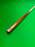 A close-up of a snooker cue shaft with a richly figured brown wood finish, resting diagonally on a green snooker table felt.