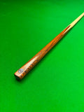 A close-up of a ash snooker cue shaft resting diagonally on a green felt snooker table surface. The handle is made of polished wood with a dark grain pattern and a small circular Pro Edge logo near the butt, while the blank is a light natural wood tone.