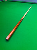 A wooden snooker cue with a glossy orange-brown shaft and a light cream tip, resting diagonally on a green felt snooker table.