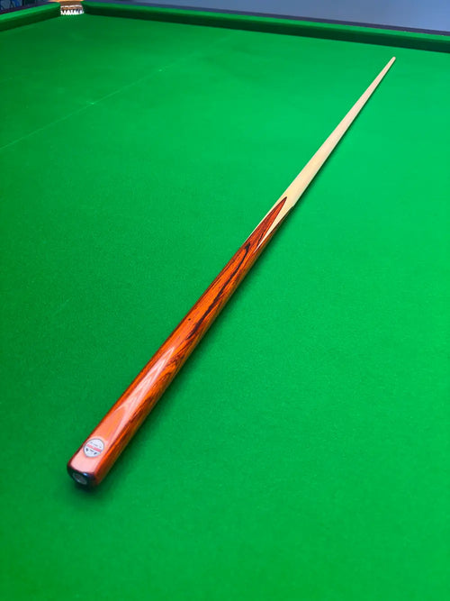 A wooden snooker cue with a glossy orange-brown shaft and a light cream tip, resting diagonally on a green felt snooker table.