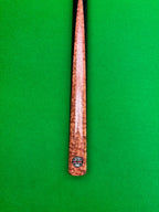 A close-up vertical shot of a snooker cue shaft with a prominent bubinga burl splice, resting on a green felt table surface. A circular Pro Edge brand label is visible near the butt end.