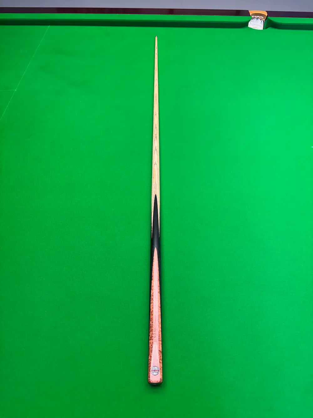 A single snooker cue laid vertically on a bright green snooker table, with the tip pointing upward and the butt near the bottom edge.