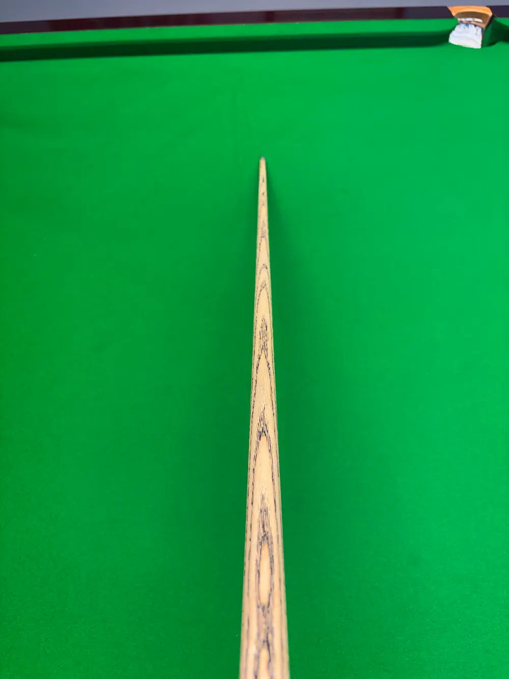 Close-up of a wooden snooker cue shaft pointing vertically down the center of a green snooker table, with the pocket area and chalk visible in the top right corner.