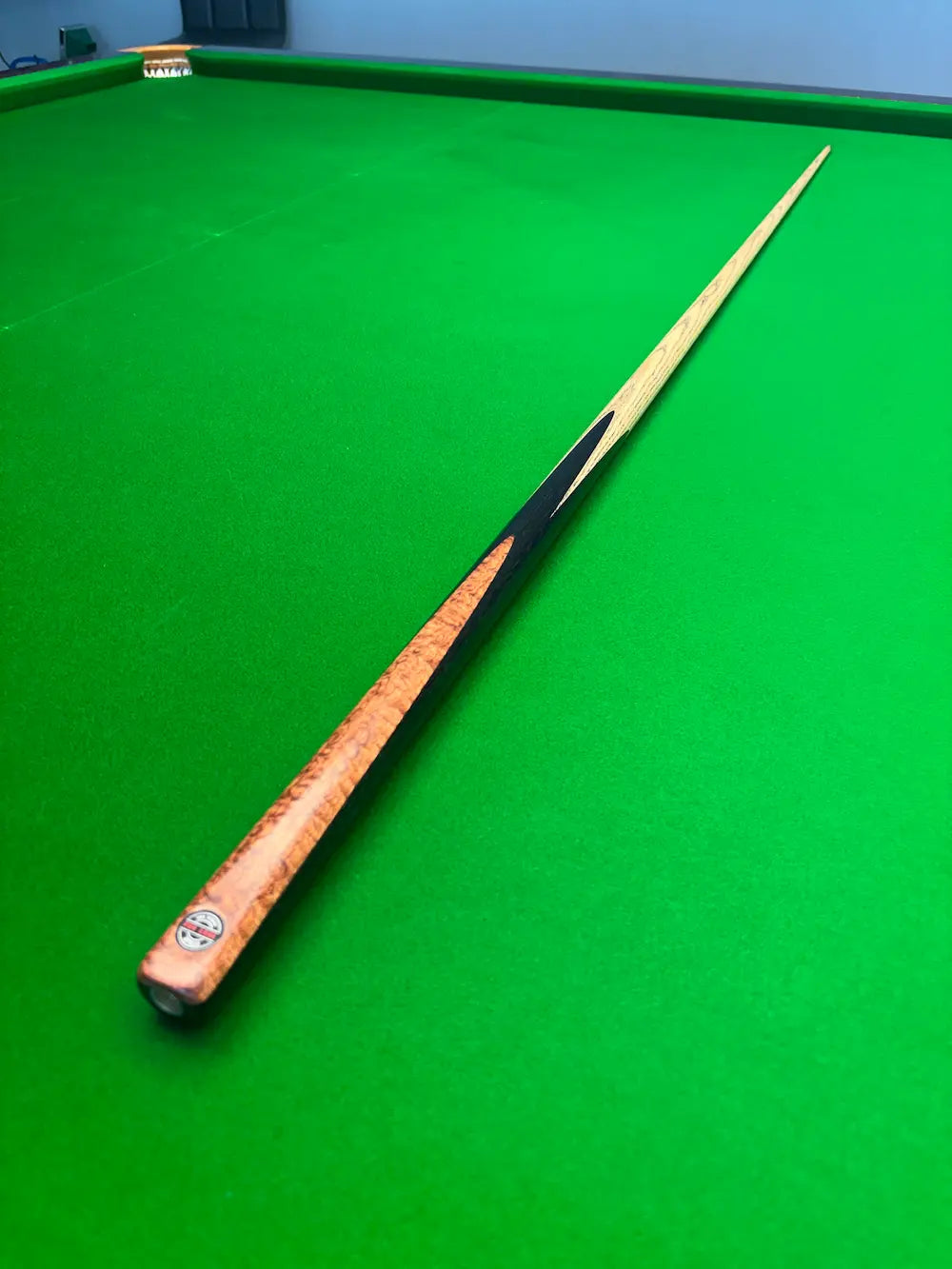 A wooden snooker cue resting diagonally on a green snooker table, showing a natural wood finish with a black painted section near the tip.