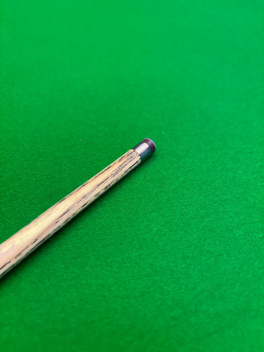 A close-up of a wooden cue with a silver ferrule and dark tip, resting diagonally on a bright green felt surface.