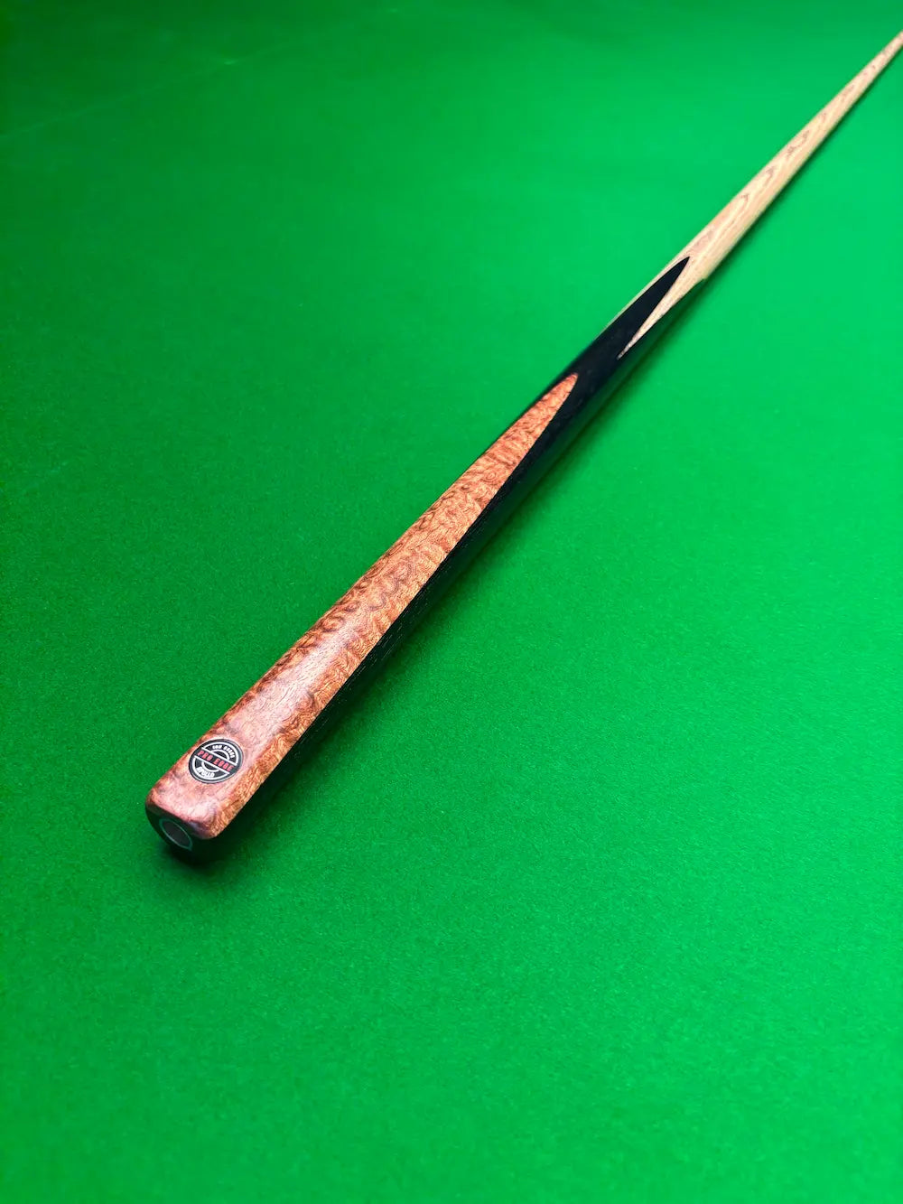 A close-up of a wooden snooker cue shaft resting diagonally on a green felt table surface, showing the wood grain.