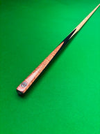 A close-up of a wooden snooker cue shaft resting diagonally on a green felt table surface, showing the wood grain.