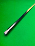 A close-up of a snooker cue shaft with a black handle and natural wood tip, resting diagonally on a green felt surface.