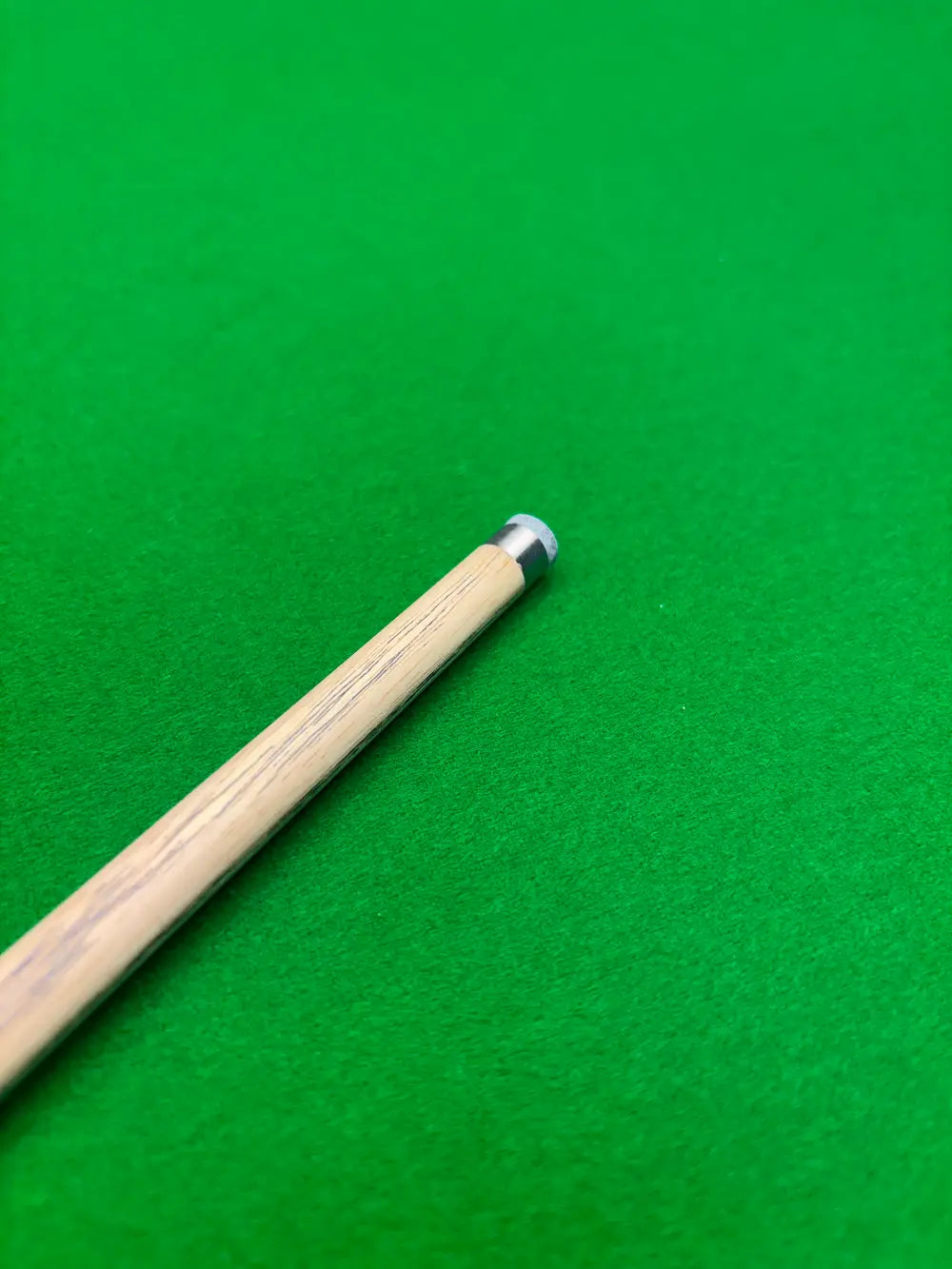 Close-up of a wooden snooker cue shaft with a silver and black tip, resting diagonally on a green felt surface.