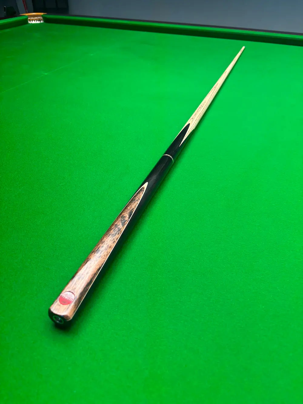 A single snooker cue resting diagonally on a green cloth snooker table, with the butt cap showing a red and white logo and light wood shafts.