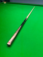 A single snooker cue resting diagonally on a green cloth snooker table, with the butt cap showing a red and white logo and light wood shafts.