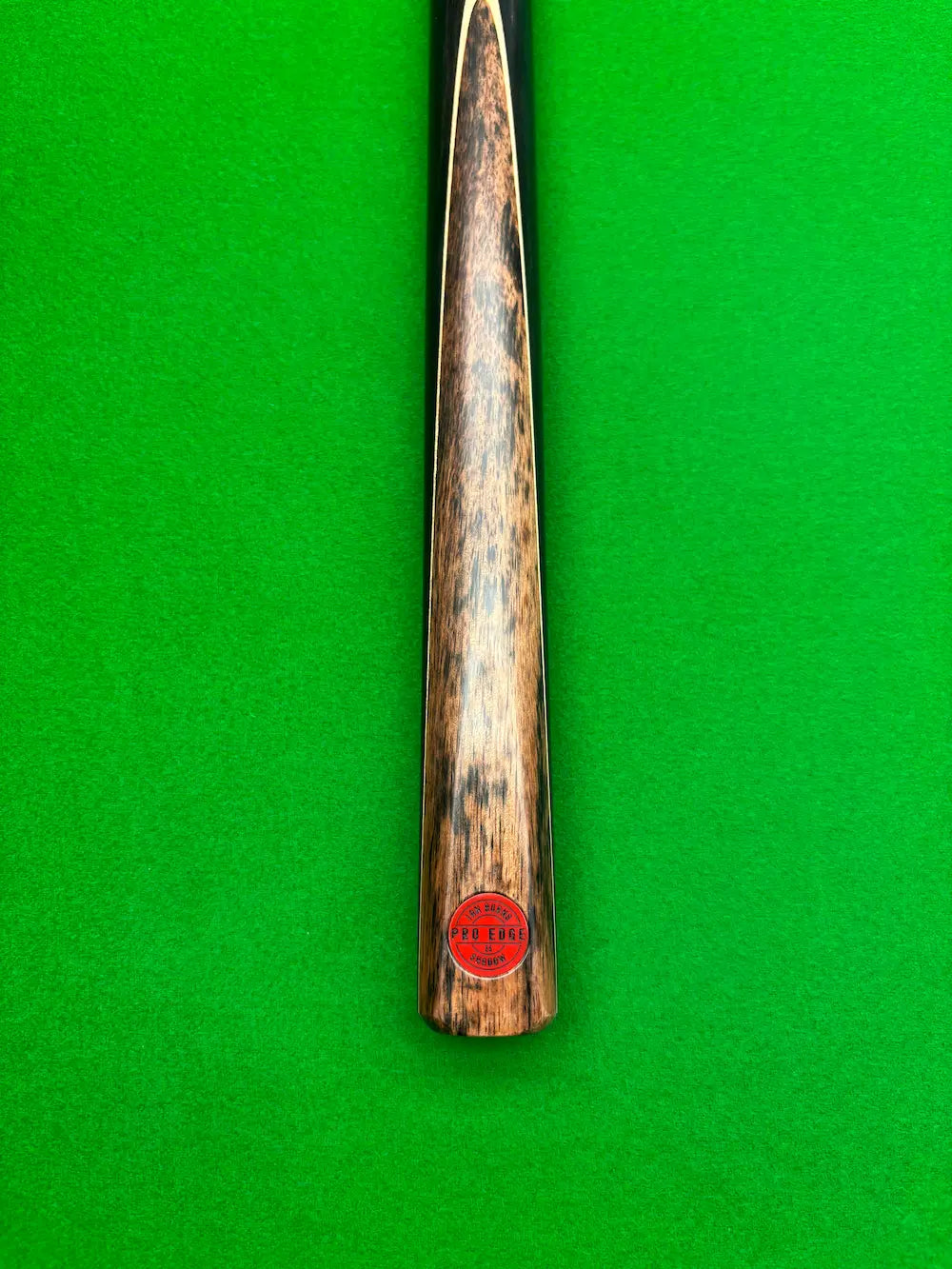 Close-up of a wooden snooker cue shaft with a red circular 'Pro Edge' brand sticker, set vertically against a bright green felt background.