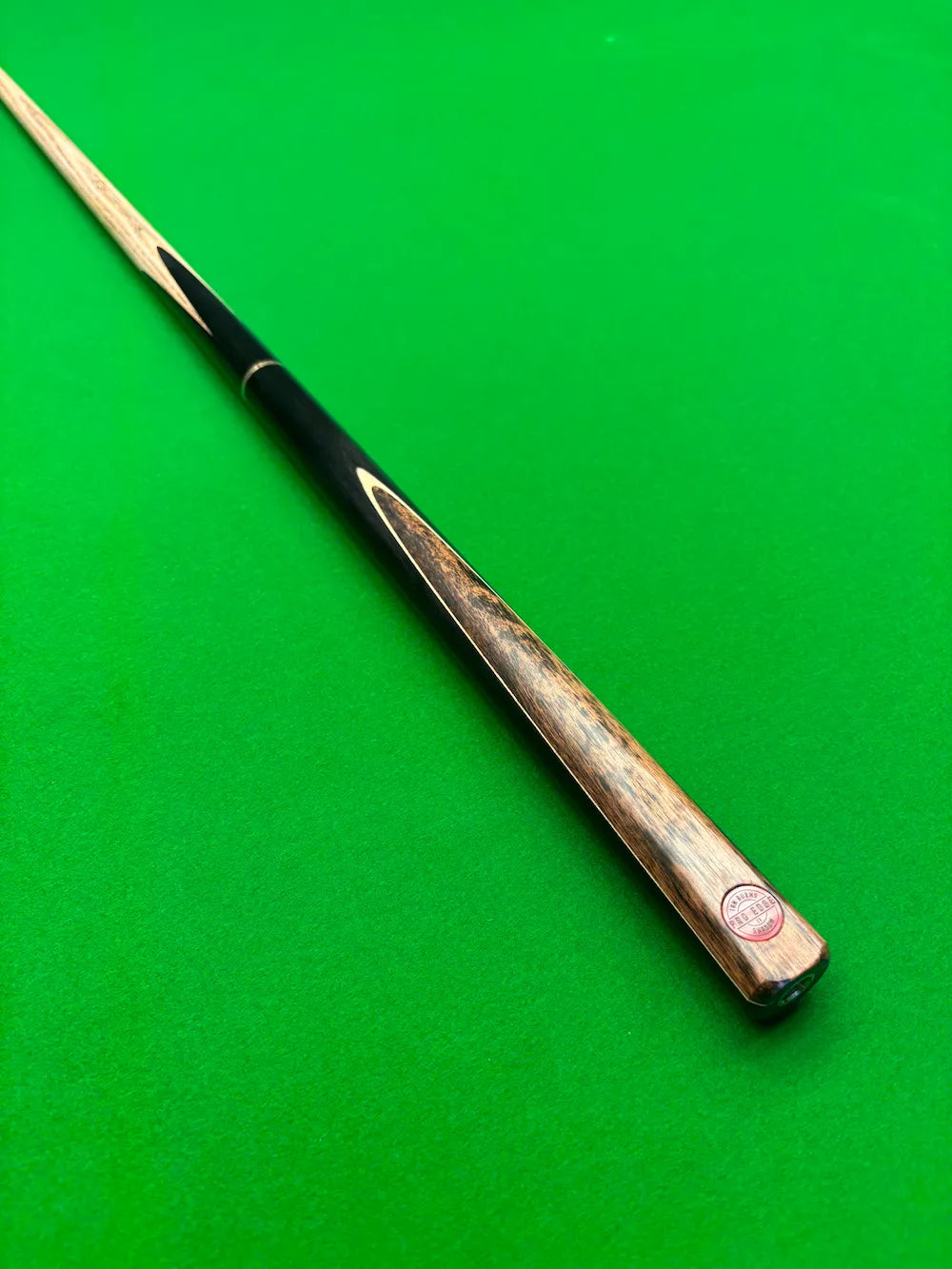 A close-up of a snooker cue shaft with a black grip and a natural wood butt cap, resting diagonally on a green snooker table felt.