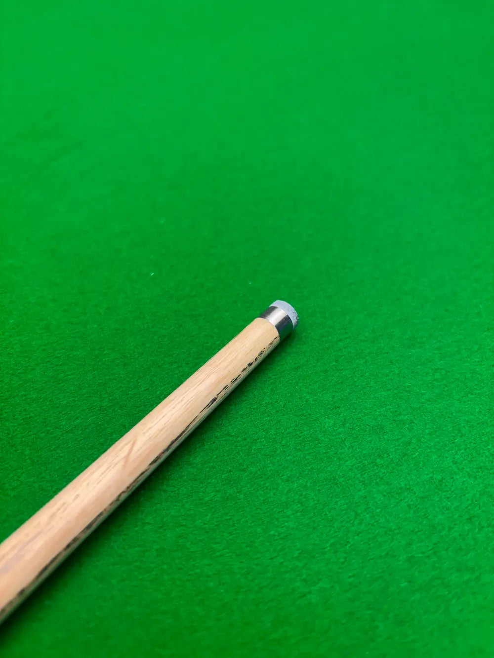 Close-up of a wooden snooker cue shaft with a silver ferrule, resting diagonally on a green felt surface.