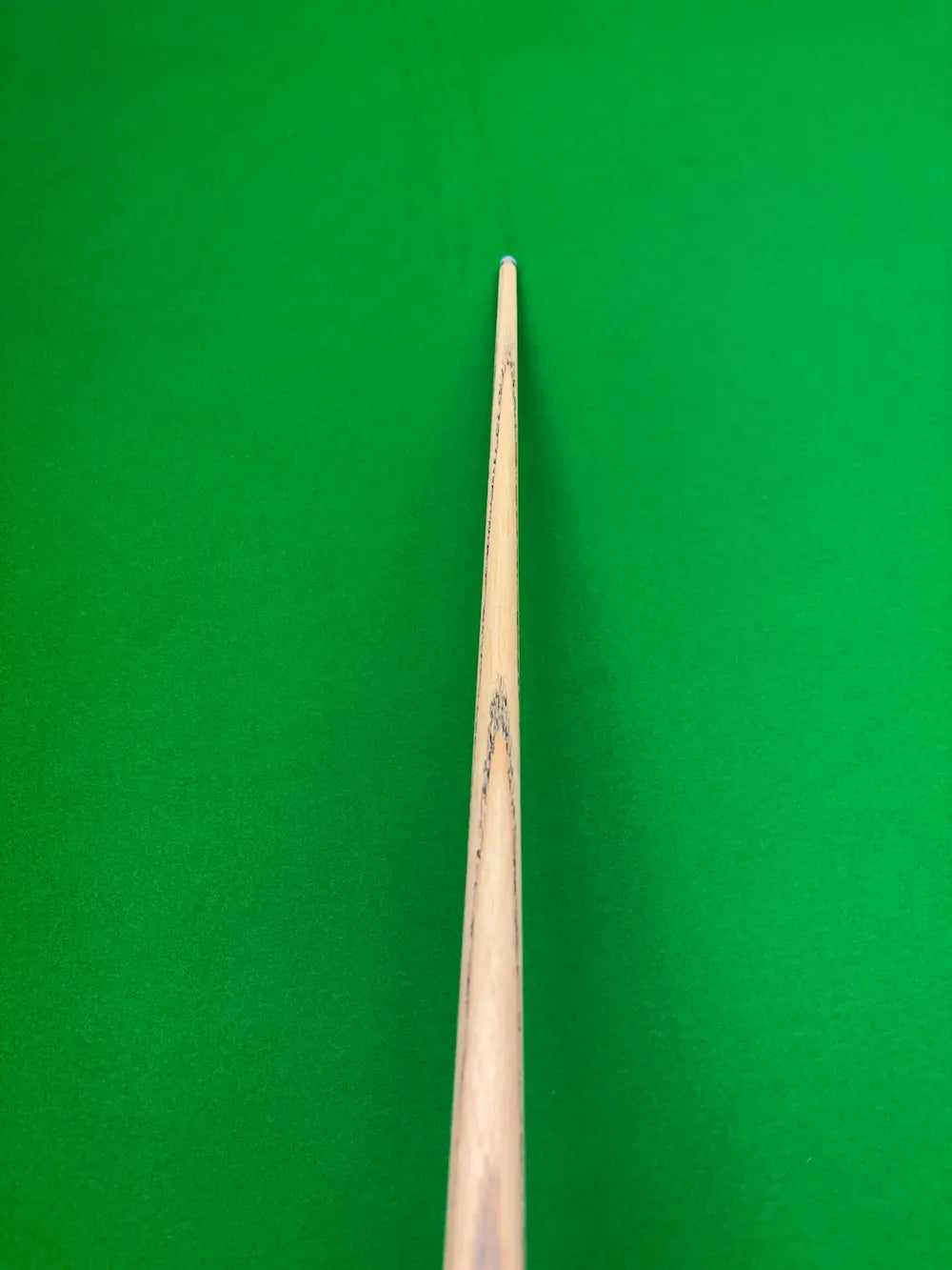A close-up vertical shot of a wooden snooker cue shaft against a bright green felt background.