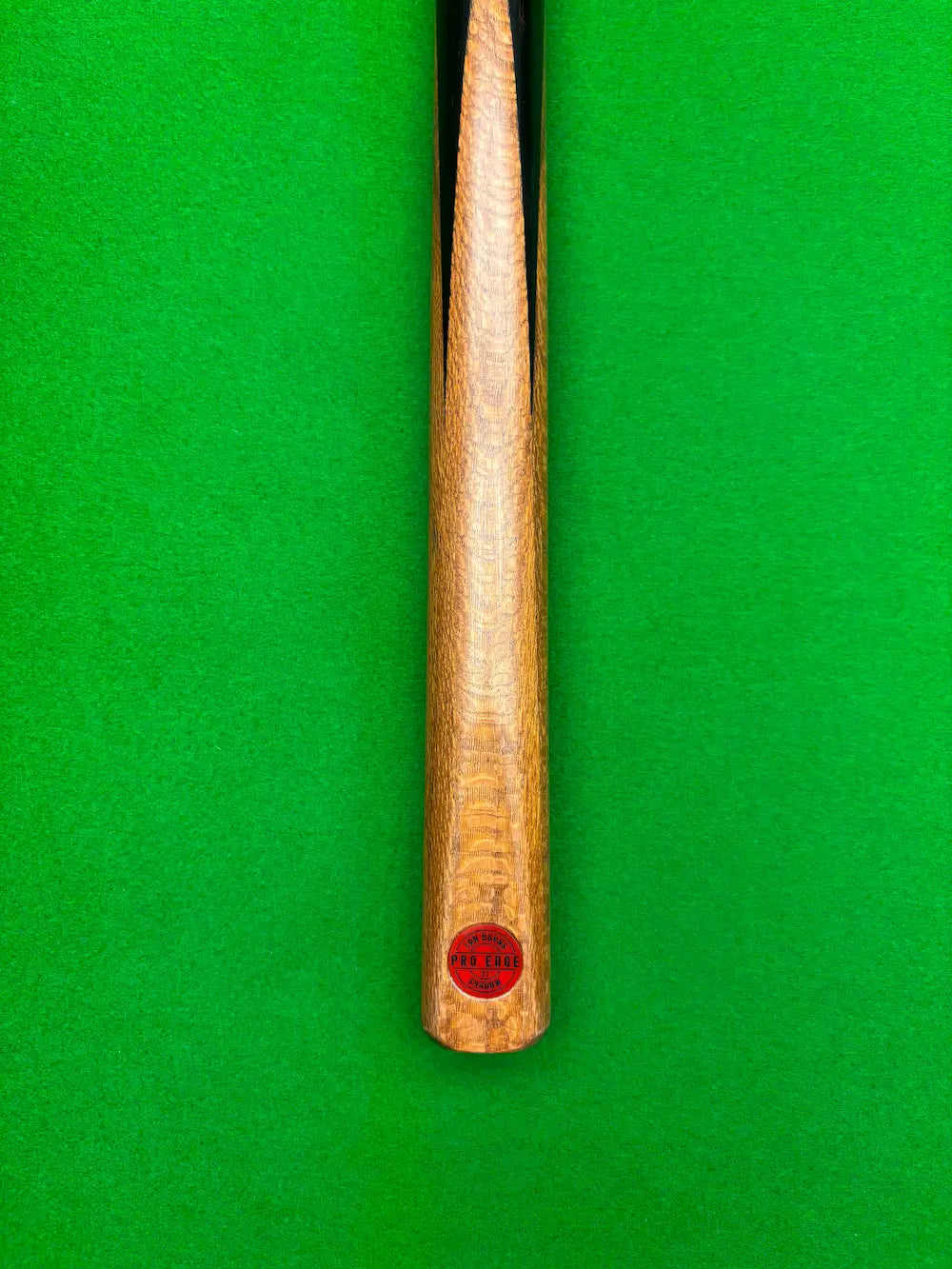 Close-up of a wooden snooker cue shaft with a red circular 'Pro Edge' brand badge, laying on a snooker table