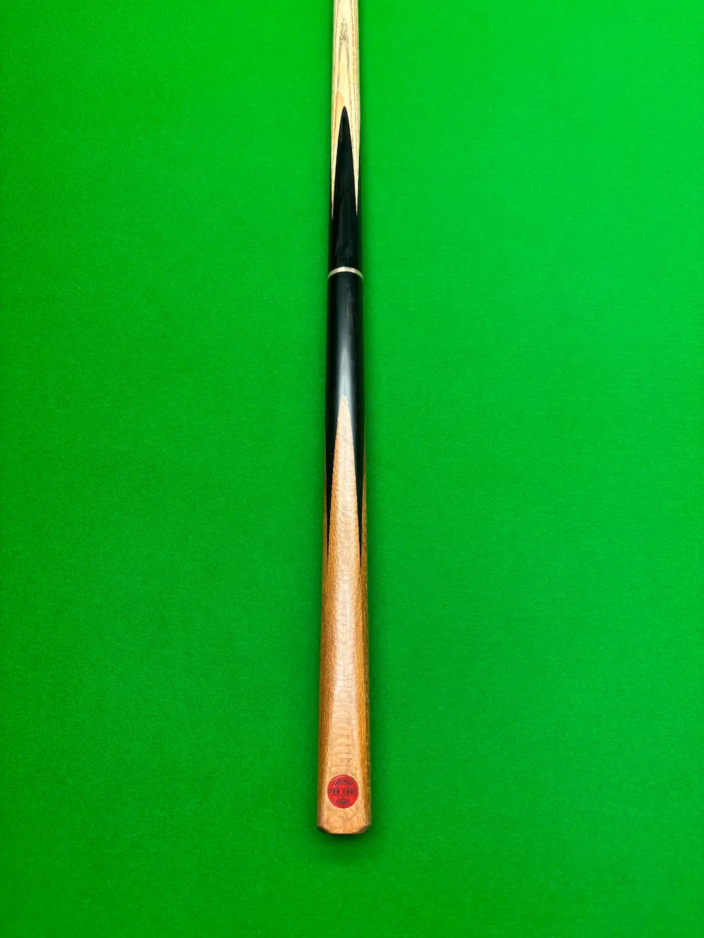 A close-up of a snooker cue shaft with a black and light wood striped finish and a red circular logo near the butt, set against a solid green felt background.