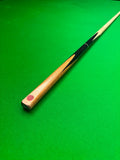 A wooden snooker cue resting diagonally on a green felt table surface, showing a light wood shaft, black painted sections, and a red cue tip.