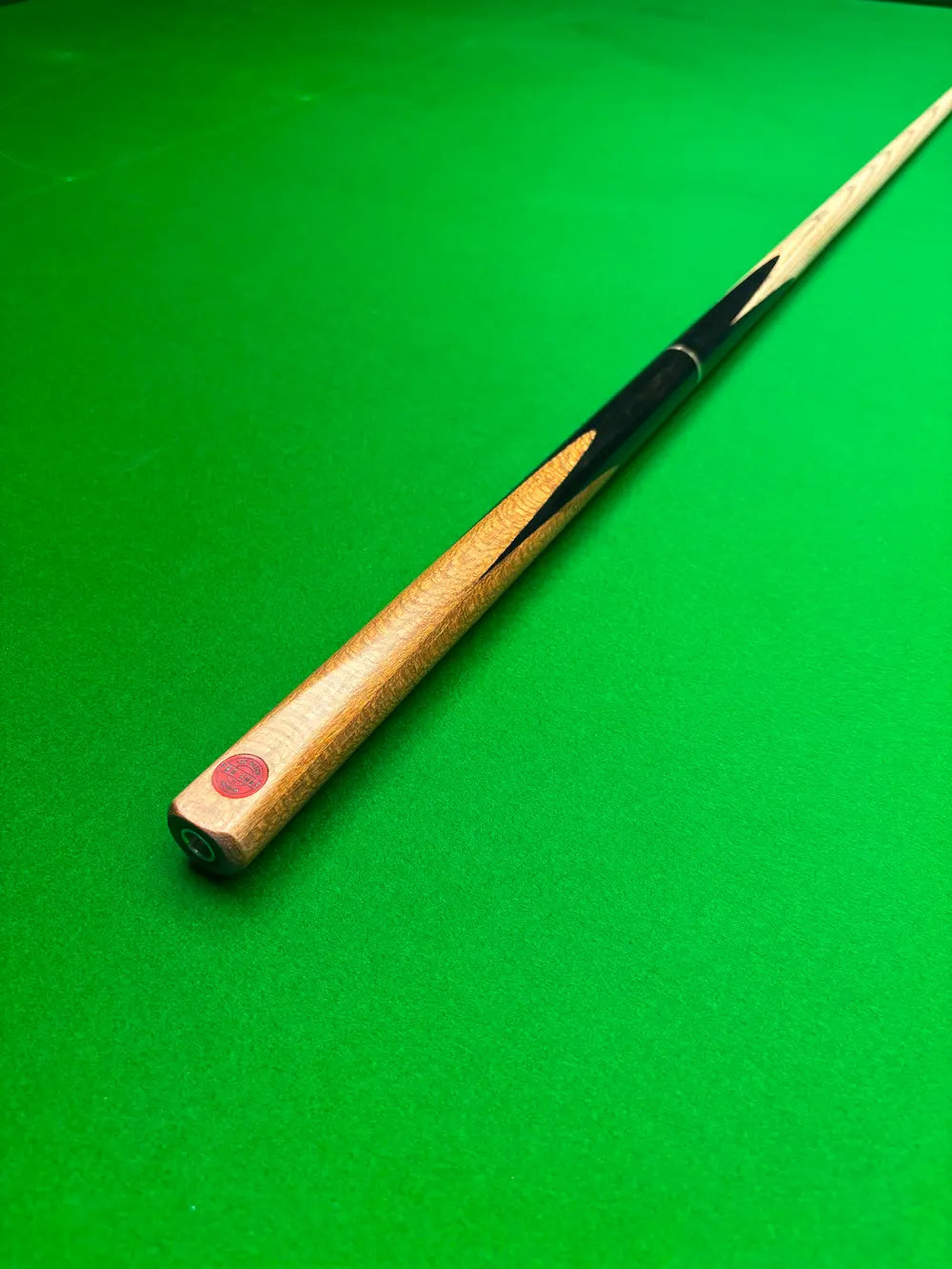 A wooden snooker cue resting diagonally on a green felt table surface, showing a light wood shaft, black painted sections, and a red cue tip.