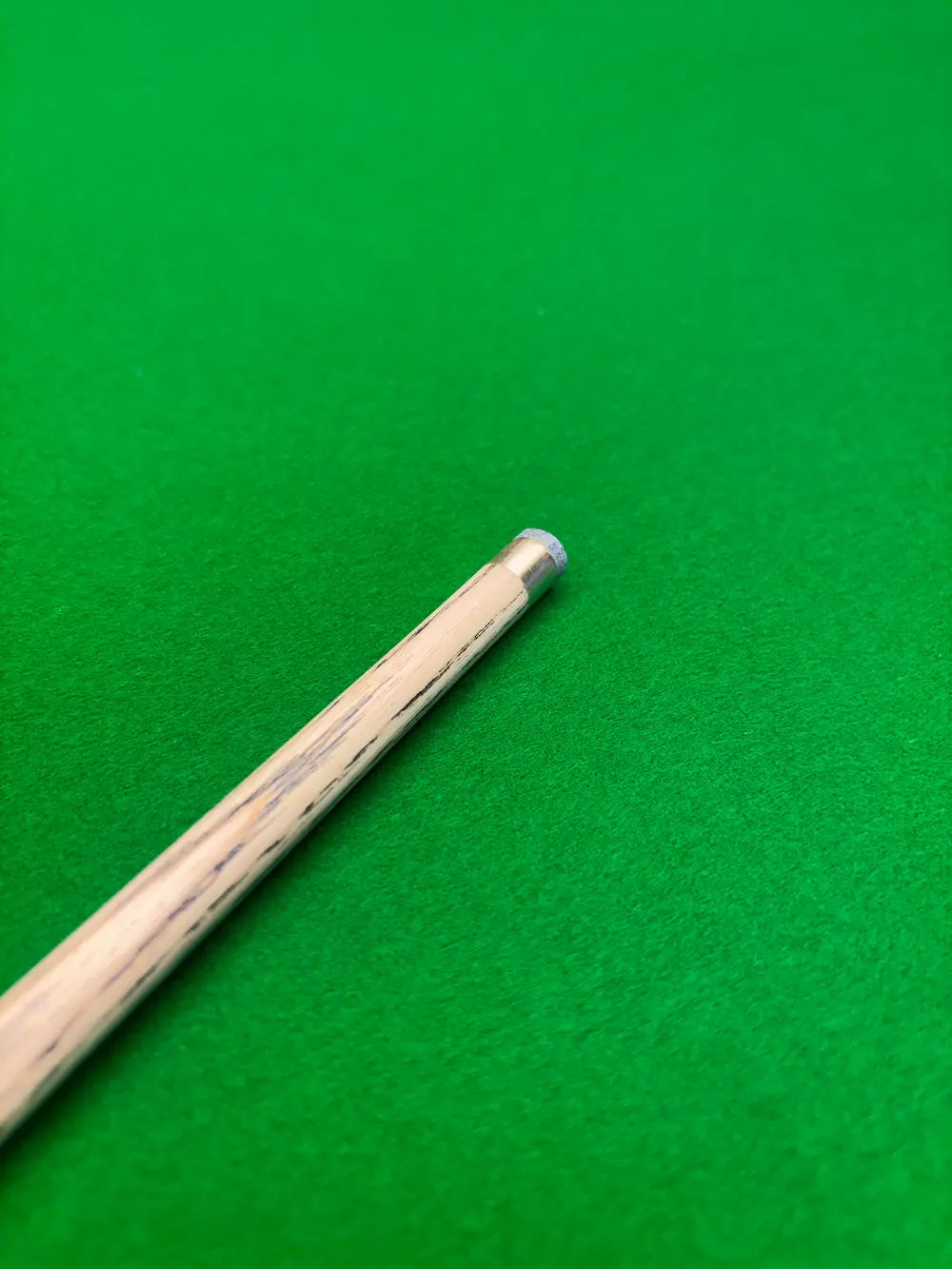 Close-up of a wooden snooker cue shaft with a silver metal tip resting on a green felt surface.