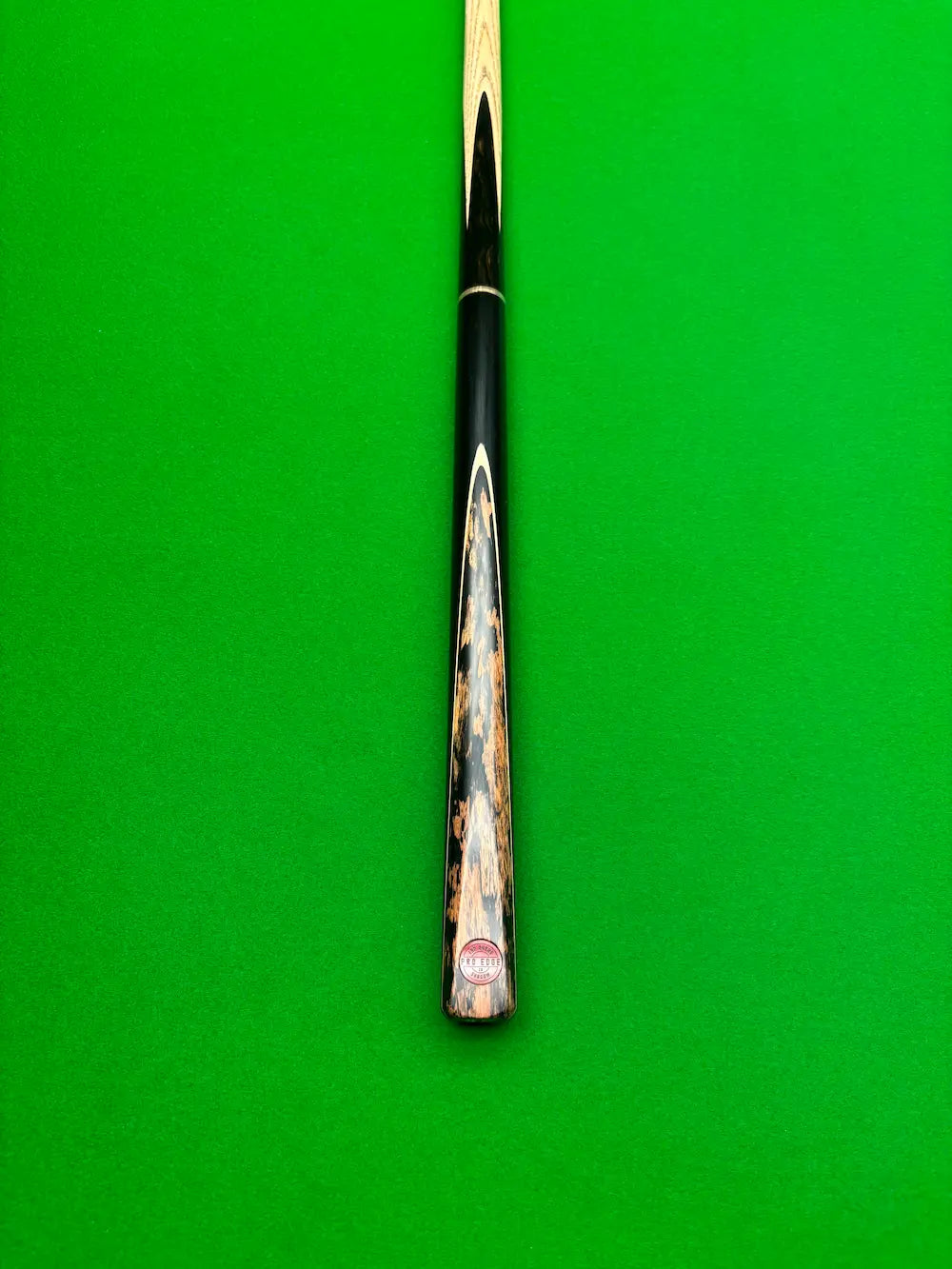 A close-up of a snooker cue shaft resting vertically on a green felt table surface, showing the black ferrule, a decorative patterned back, and the butt cap.