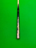 A close-up of a snooker cue shaft resting vertically on a green felt table surface, showing the black ferrule, a decorative patterned back, and the butt cap.