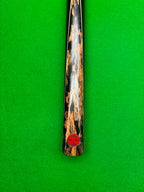 A close-up of a snooker cue shaft with a distinctive black and brown mottled wood pattern, resting vertically on a green felt surface. A red circular sticker is visible near the bottom of the shaft.