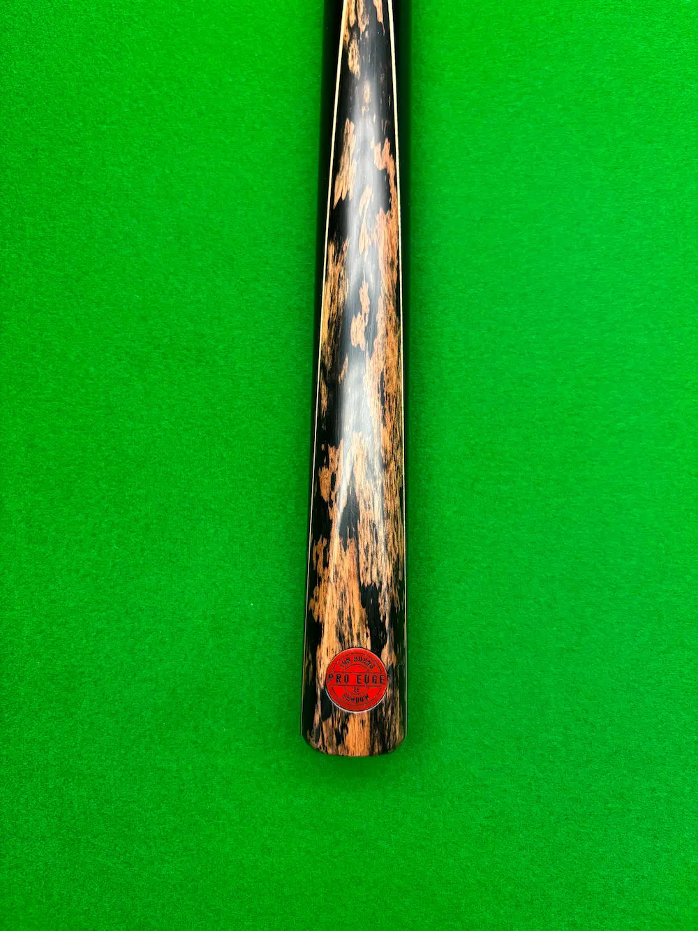 A close-up of a snooker cue shaft with a distinctive black and brown mottled wood pattern, resting vertically on a green felt surface. A red circular sticker is visible near the bottom of the shaft.