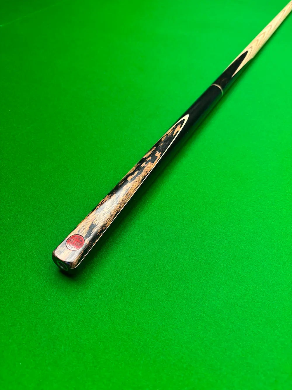 A close-up of a snooker cue shaft resting diagonally on a vibrant green felt table surface, showing the polished wood end with a red brand stamp.