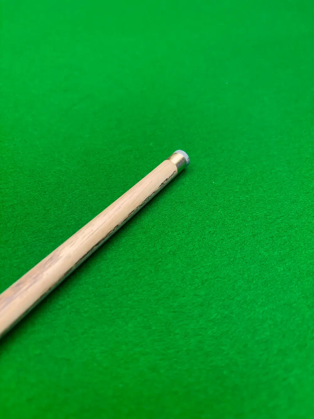A close-up of a wooden snooker cue shaft with a silver-colored ferrule, resting diagonally on a vibrant green felt surface.