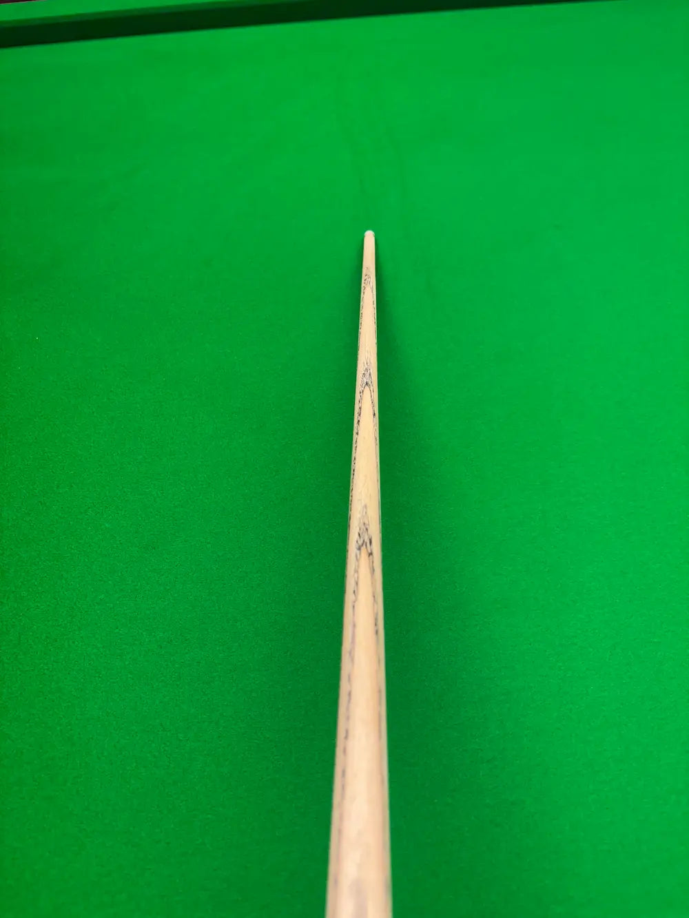 A close-up shot of a wooden snooker cue shaft pointing toward a green felt snooker table surface.