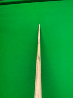 A close-up shot of a wooden snooker cue shaft pointing toward a green felt snooker table surface.