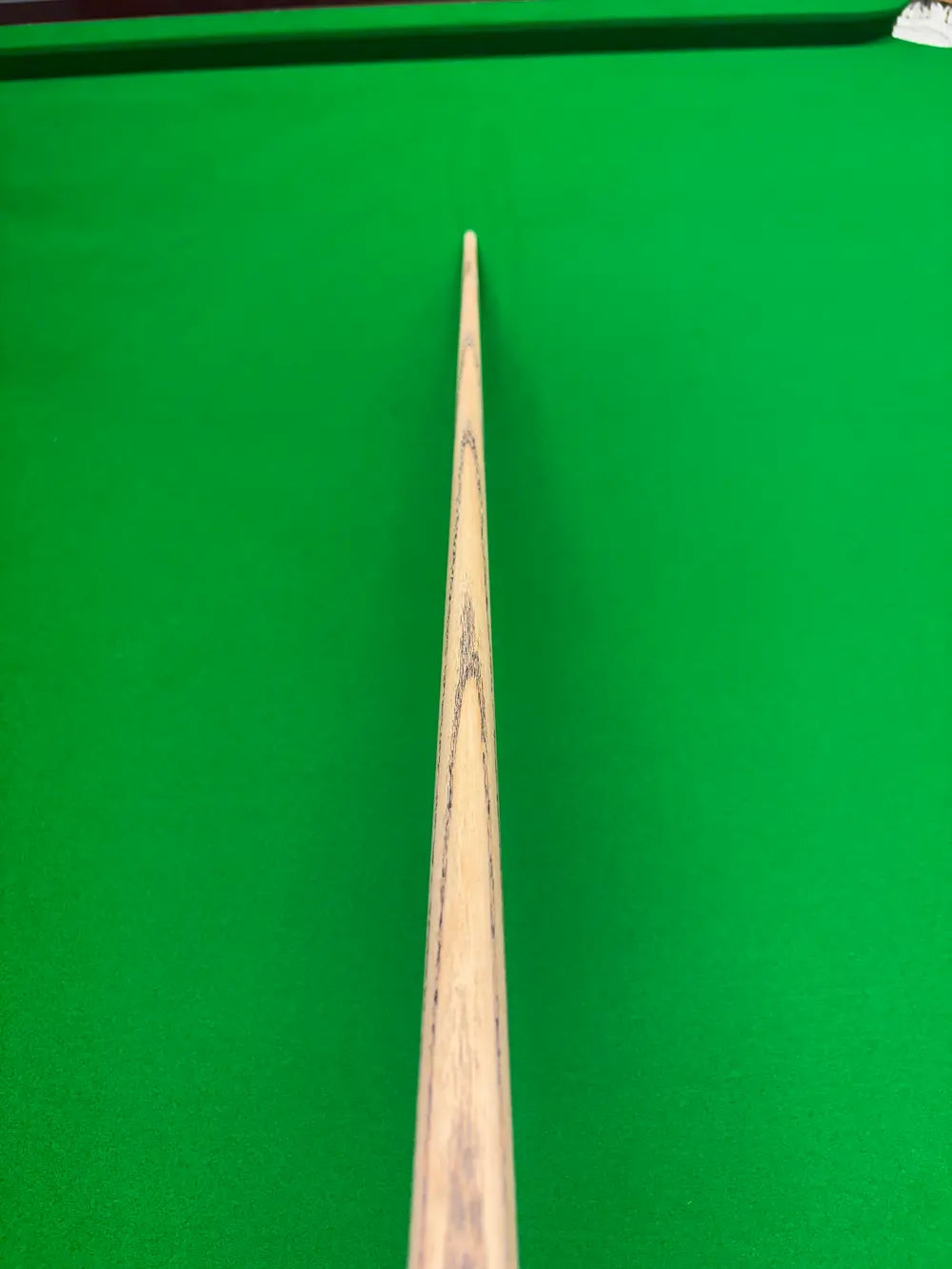 A close-up, top-down view of a wooden snooker cue shaft positioned vertically against a bright green snooker table felt background.