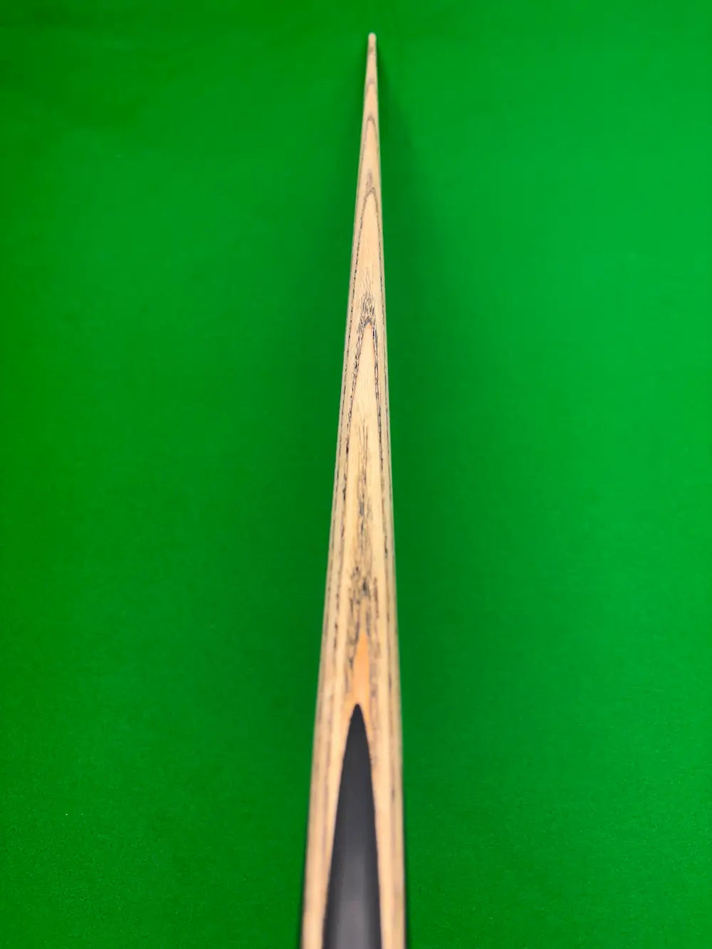 Close-up of a wooden snooker cue shaft with a black ferrule, positioned vertically against a bright green snooker table felt background.