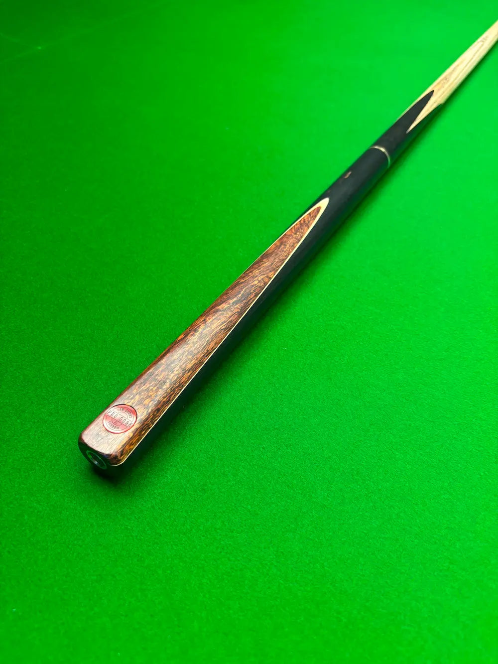 A close-up of a snooker cue shaft with a dark black handle and a brown wood grain face, resting diagonally on a green felt table surface.