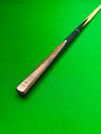 A close-up of a snooker cue shaft with a dark black handle and a brown wood grain face, resting diagonally on a green felt table surface.
