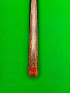 A close-up of a snooker cue shaft with a rich, swirling brown and golden wood grain finish, featuring a small red circular stamp near the butt, set against a vibrant green snooker felt background.