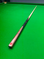 A wooden snooker cue with a black grip and red tip, resting diagonally on a green snooker table.