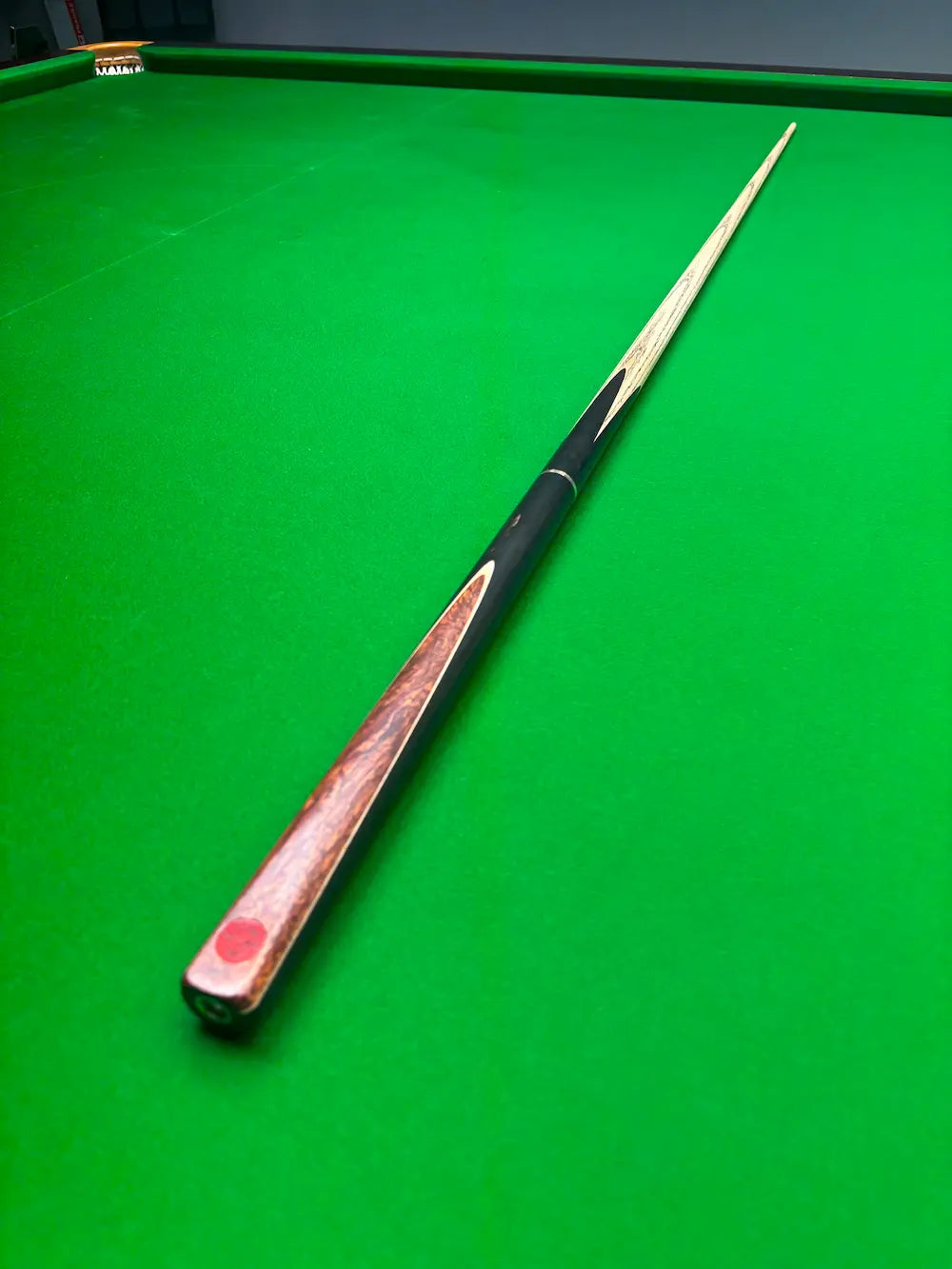 A wooden snooker cue with a black grip and red tip, resting diagonally on a green snooker table.