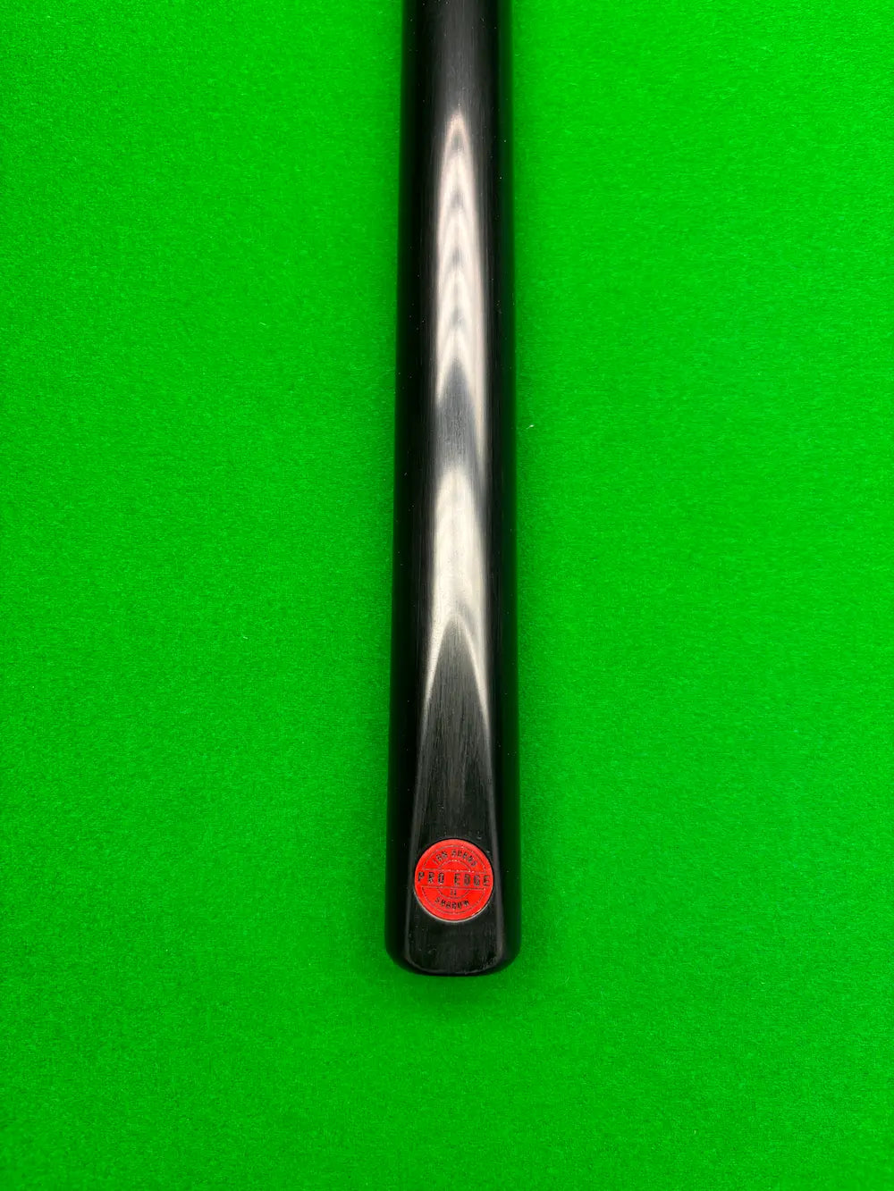 Close-up of a black snooker cue shaft with a red circular Pro Edge' brand sticker, positioned vertically against a bright green snooker felt background.