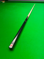 A single snooker cue with a glossy black handle and light wood shaft resting diagonally on a green felt snooker table. A red circular Pro Edge logo is visible on the end cap of the handle.