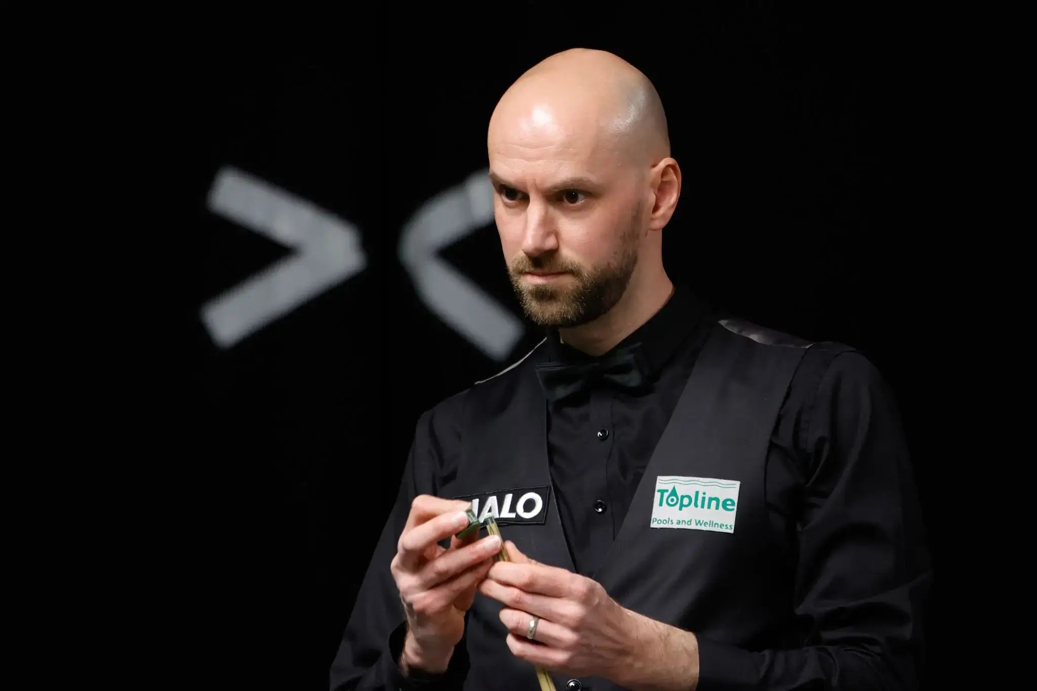 Man in a black shirt with logos holding a snooker cue against a dark background